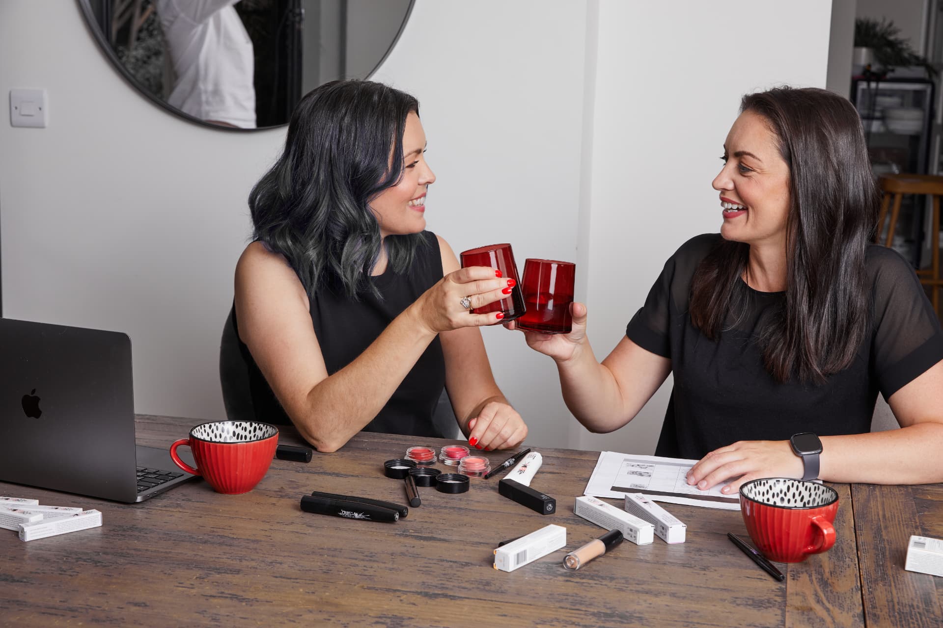 Sarah and Emma, the sisters behind CODE Beautiful, laughing together over a table of makeup